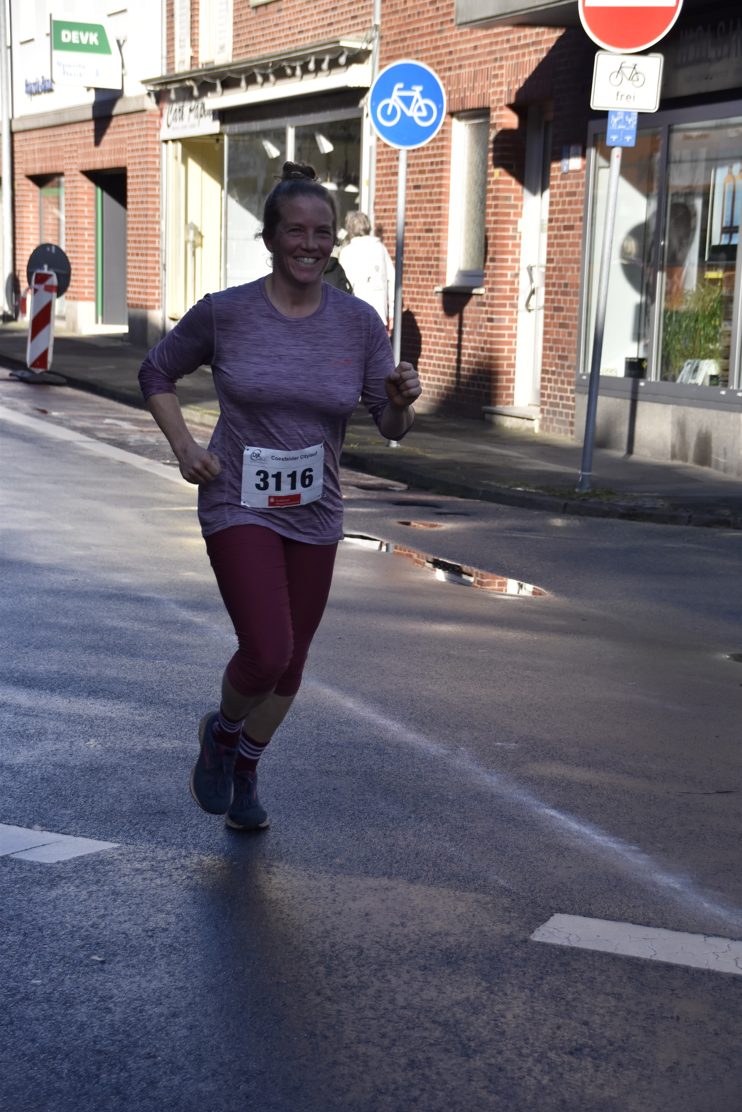 Läuferin in sportlicher Kleidung mit Startnummer 3116 joggt durch eine Straßenkulisse mit Geschäften und Verkehrsschildern.