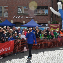 Ein Moderator in blauer Jacke spricht mit Publikum, umgeben von Zuschauern und bunten Ständen auf einem Fest.