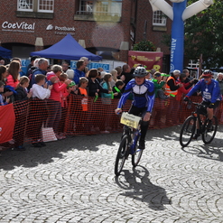 Zwei Radfahrer auf einer gepflasterten Straße fahren einem jubelnden Publikum entgegen, während sie ein Rennen beenden.
