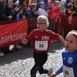 Zwei Kinder in Sportkleidung laufen beim Coesfelder Citylauf, viele Zuschauer stehen applaudierend im Hintergrund.