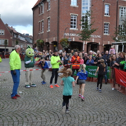 Zwei Kinder laufen im Zielbereich eines Wettlaufs, umgeben von jubelnden Zuschauern und Helfern in Neonkleidung.