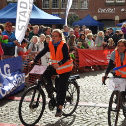 Zwei Radfahrerinnen in auffälligen Westen fahren durch das Ziel eines Radrennens, umgeben von jubelndem Publikum.