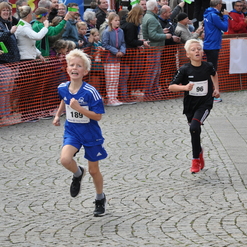 Zwei Kinder im Wettlauf, eines in blauem und das andere in schwarzem Trikot, laufen auf einem gepflasterten Platz.
