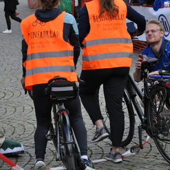 Zwei Personen in leuchtend orangefarbenen Westen stehen mit Fahrrädern vor einer Menschenmenge auf einem Platz.