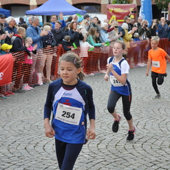 Drei Kinder laufen beim Wettlauf, umgeben von jubelnden Zuschauern und einem roten Sparkassen-Banner.