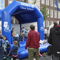 Kinder spielen auf einer blauen Hüpfburg, umgeben von Erwachsenen, in einem belebten Stadtbereich.