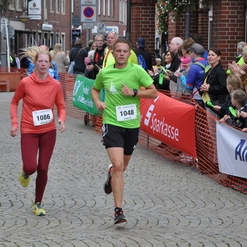 Zwei Läufer, einer in Grün und einer in Rot, laufen auf einer Straße, umgeben von jubelnden Zuschauern.