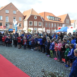 Gruppierung von Kindern und Erwachsenen bei einer Veranstaltung auf einem Platz, Blick auf eine Bühne im Vordergrund.