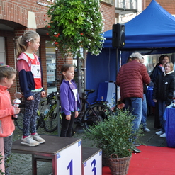 Drei Mädchen stehen auf dem Siegertreppchen bei einer Sportveranstaltung, während ein Mann ihnen eine Urkunde überreicht.