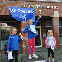 Drei Mädchen stehen auf dem Siegertreppchen, feiern ihre Platzierungen mit Pokalen und einer Urkunde vor einem Gebäude.