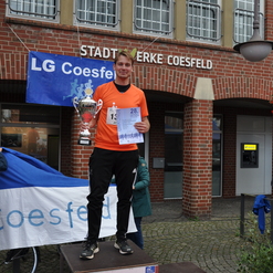 Mann in orangeem Shirt steht auf dem Siegerpodest mit Pokal und Urkunde vor der Gebäude-Fassade in Coesfeld.