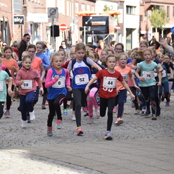 Gruppe junger Mädchen in Sportbekleidung beim Start eines Rennen auf einer gepflasterten Straße, Zuschauer im Hintergrund.
