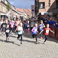Gruppe von Mädchen in Sportkleidung bei einem Laufereignis auf einer gepflasterten Straße, umgeben von Zuschauern.