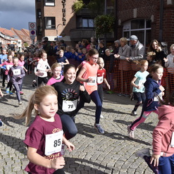 Eine Gruppe von Kindern läuft fröhlich einen Wettlauf auf einer befestigten Straße, Zuschauer jubeln entlang der Strecke.