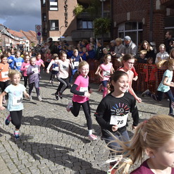 Mädchen in Sportkleidung laufen begeistert bei einem Kinderlauf durch eine belebte Straße mit Zuschauern.