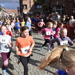 Gruppe von Mädchen läuft fröhlich einen Straßenlauf, umgeben von Zuschauern auf einer gepflasterten Straße.
