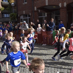 Gruppierung von Kindern beim Laufwettbewerb auf einer Straße, umgeben von Zuschauern und orangefarbener Absperrung.