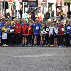 Gruppierung von Kindern in Sportbekleidung mit Startnummern an der Startlinie eines Rennens auf einer gepflasterten Straße.
