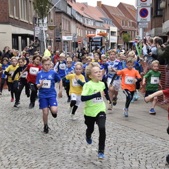 Gruppe von Kindern in bunten Lauftrikots bei einem Wettlauf auf einer gepflasterten Straße, Zuschauer beobachten.