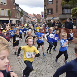 Eine Gruppe von Kindern in Sportkleidung läuft an einem Straßenschild entlang während eines Wettlaufs. Zuschauer beobachten.