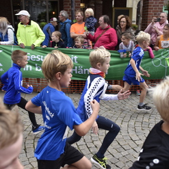 Gruppe von Kindern in blauen Trikots beim Laufen auf einer Straße, gekennzeichnet von Zuschauer*innen und einem Banner.