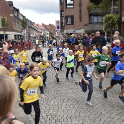 Gruppierung von Kindern in Sportbekleidung beim Start eines Straßenrennens, umgeben von Zuschauern in einer Stadt.
