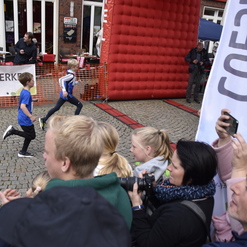 Zwei Kinderlaufen während eines Laufevents, umgeben von jubelnden Zuschauern und einer Banner.