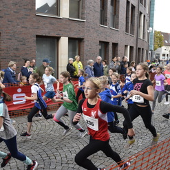 Gruppe von Mädchen in Sportkleidung beim Laufen in einer Stadt, Zuschauer stehen hinter einem Absperrgitter.