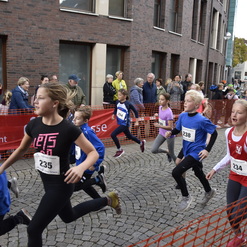 Gruppe von Kindern läuft im Wettkampf auf einer Steinpflasterstraße, umgeben von Zuschauern hinter einer Absperrung.