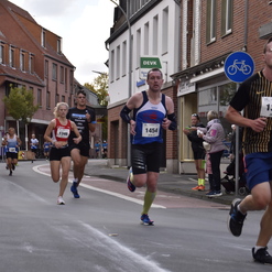 Vier Läufer in Wettkleidung laufen auf einer Straße, umgeben von Zuschauern und Gebäuden im Hintergrund.