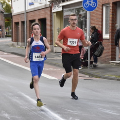 Verschiedene Läufer, darunter ein Junge in blauer Sportkleidung, laufen eine Straße entlang. Zuschauer stehen am Rand.