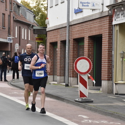 Vier Läufer in Sportkleidung joggen auf einer Stadtstraße, während Zuschauer ihnen zujubeln und Getränke reichen.