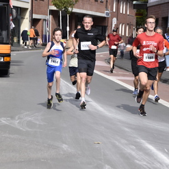 Vier Läufer in Sportkleidung nehmen an einem Straßenlauf teil; zuschauende Menschen und ein Bus im Hintergrund.