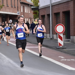 Läufer in Sportoutfits sprinten auf einer Straße, während Zuschauer am Rand anfeuern. Verkehrsschild im Hintergrund sichtbar.