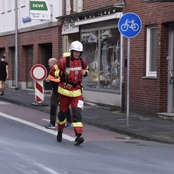 Ein Feuerwehrmann in voller Ausrüstung joggt auf einer Straße, während Passanten und Schilder im Hintergrund zu sehen sind.