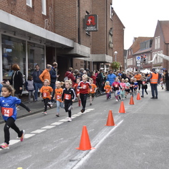Kinder in bunten T-Shirts laufen einen Wettlauf auf einer Straße, umgeben von Zuschauern und orangefarbenen Pylonen.