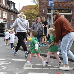 Gruppen von Kindern und Erwachsenen laufen gemeinsam auf einer Straßenkreuzung, umgeben von einheimischen Geschäften.
