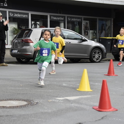 Kinder laufen beim Wettlauf durch einen markierten Streckenabschnitt mit Pylonen und jubelnden Zuschauern im Hintergrund.