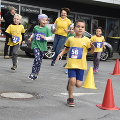 Kinder in Laufshirts nehmen an einem Wettlauf teil, während Zuschauer am Rand anfeuern. Mehrere Verkehrshütchen stehen bereit.