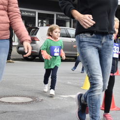 Kinder in Sportbekleidung laufen an einer Straße entlang, während Erwachsene im Hintergrund vorbeigehen.