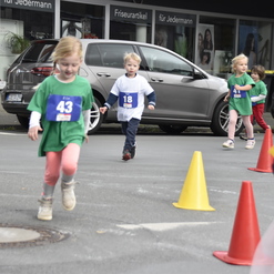 Kinder in Sportoberteilen laufen einen Wettlauf auf der Straße zwischen Pylonen, applaudierende Zuschauer im Hintergrund.
