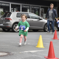 Ein Mädchen in grünem T-Shirt läuft fröhlich um orangefarbene Pylonen, während Erwachsene im Hintergrund stehen.