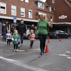 Kinder und Erwachsene laufen gemeinsam auf einer Straße, umgeben von orangefarbenen Pylonen und Zuschauern.