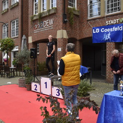 Frau auf dem Siegerpodest lächelt, während eine Gruppe Zuschauer und ein Redner ihr Anerkennung zollen.