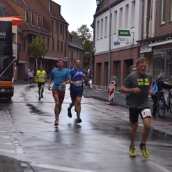 Teilnehmer eines Straßenlaufs laufen in einer nassen, schmalen Straße zwischen Häusern und verkehrsbedingt abgestellten Fahrzeugen.