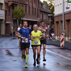 Vier Läufer in Sportbekleidung laufen auf einer nassen Straße zwischen Gebäuden während eines Wettkampfs.