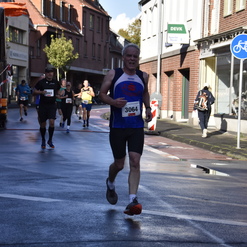 Läufer im blauen Shirt überquert eine nasse Straße während eines Wettkampfs, weitere Teilnehmer im Hintergrund.