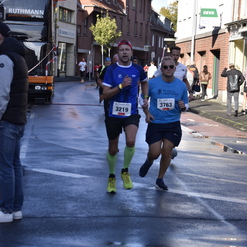 Zwei Läufer in Sportbekleidung joggen auf einer nassen Straße, vorbei an Zuschauern und einem Lkw.