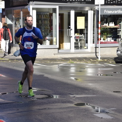 Läufer in blauem Shirt mit Startnummer 3576 joggt auf nasser Straße vor einem Friseursalon.