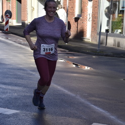 Läuferin in sportlicher Kleidung mit Startnummer 3116 joggt durch eine Straßenkulisse mit Geschäften und Verkehrsschildern.